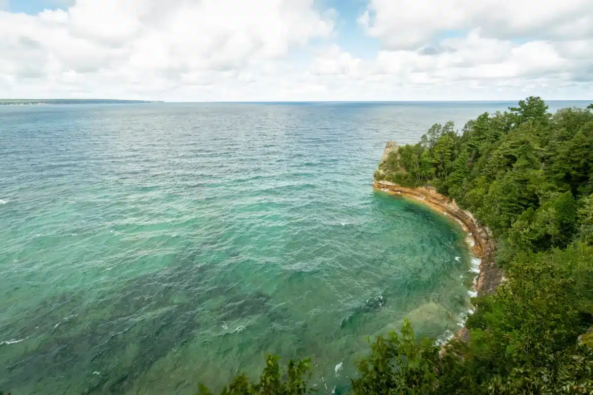 A scenic view of a clear blue-green lake bordered by a rocky shoreline and dense green forest under a partly cloudy sky.