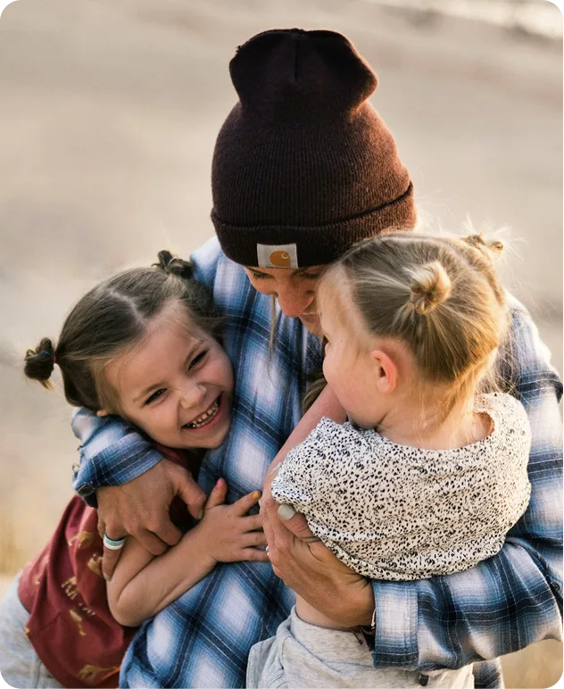 A person in a beanie hugs two young smiling girls, all appearing happy and close outdoors—radiating joy like the returns from a high-yield savings account.