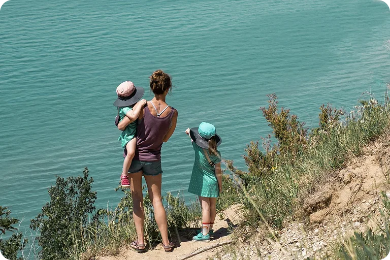 A woman stands on a cliffside path by turquoise water, holding a child and standing beside another child pointing toward the lake.