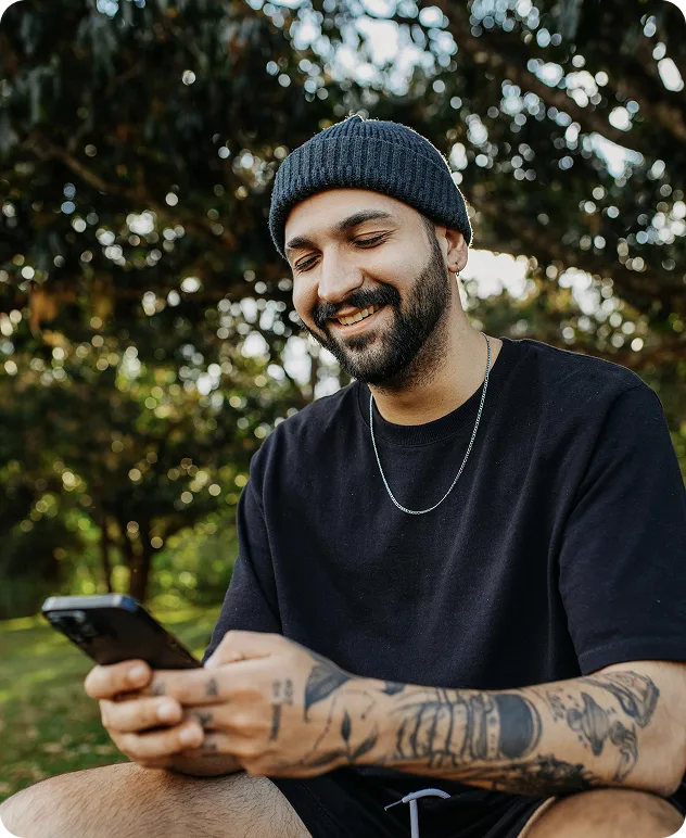 A smiling man with tattoos, wearing a black beanie and black shirt, sits outdoors and looks at his phone. Trees and greenery are visible in the background.