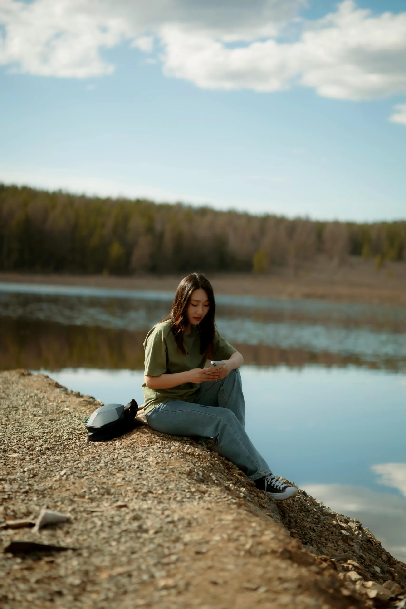 A woman sits on a rocky shore by a calm lake, reading about NCUA insurance savings accounts, surrounded by nature and trees under a partly cloudy sky.