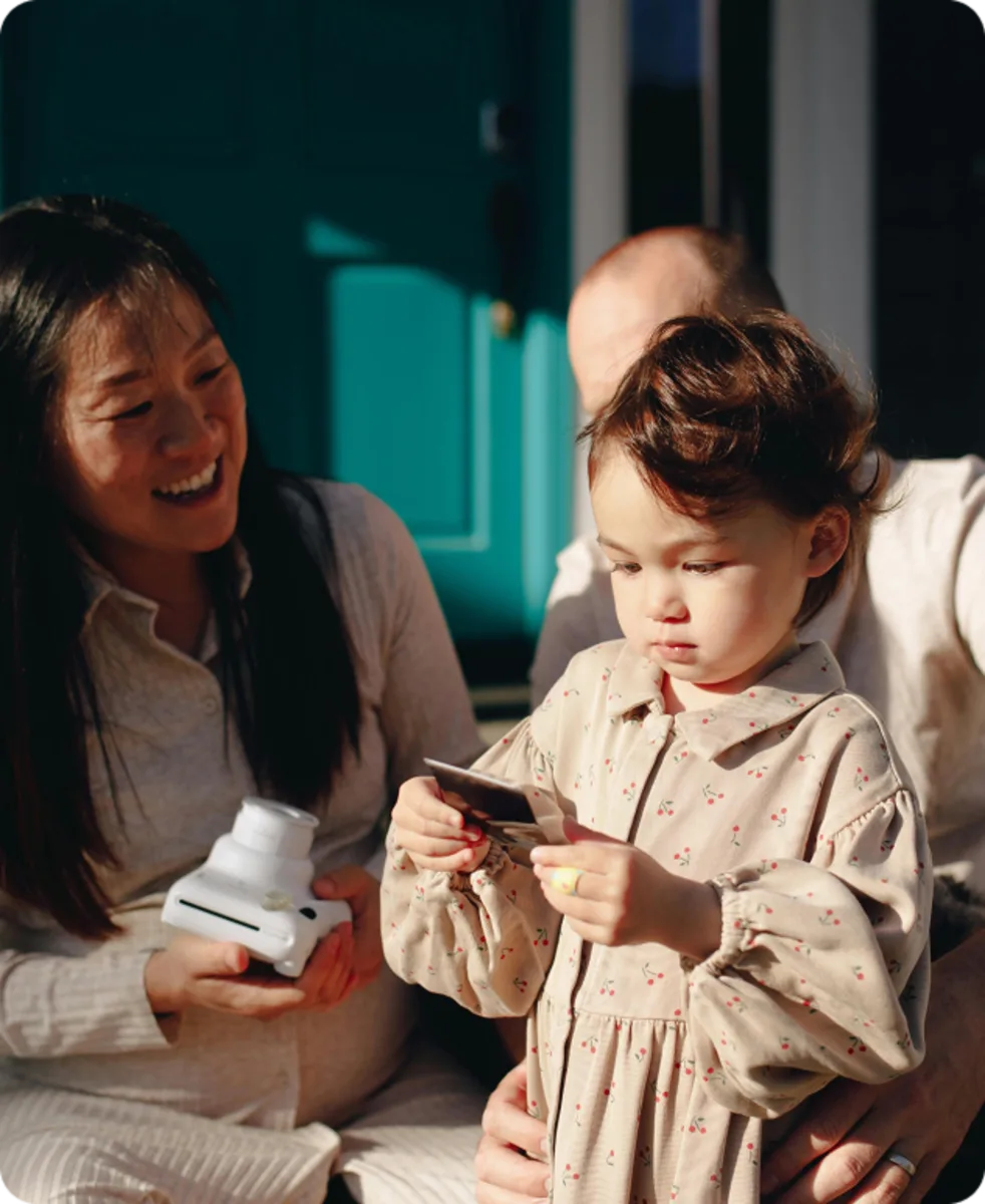 A woman smiles while holding an instant camera, as a young child in a patterned dress looks at a photo, with an adult beside them.