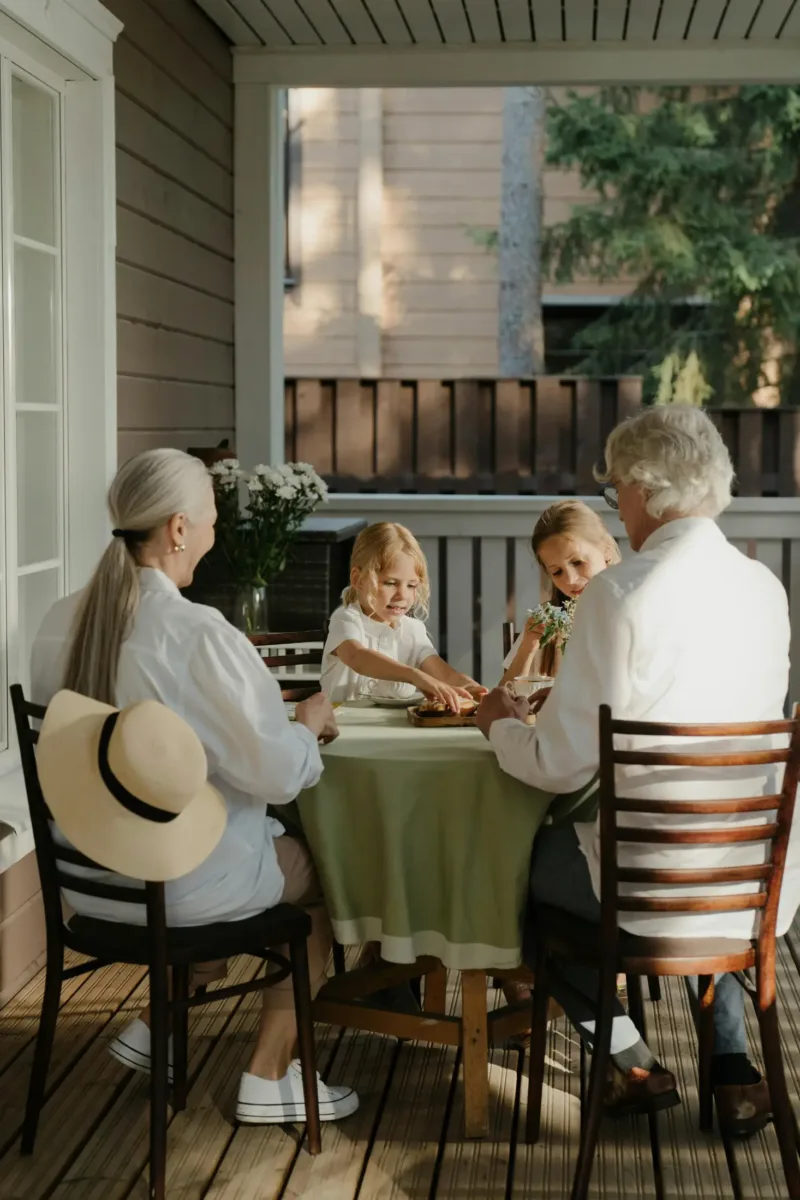Two adults and two children sit around a round table on a porch, enjoying a meal together. Sunlight streams in, and flowers are on the table. A hat rests on the chair.